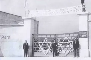 Foto in bianco e nero di tre uomini davanti a un cancello decorato con due grandi stelle di David, sotto uno striscione con scritte in ebraico; sul muro a sinistra la scritta 'Trani'