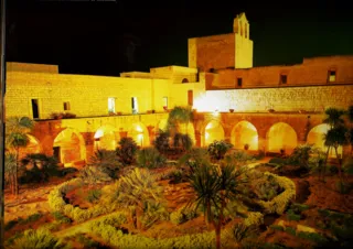 Giardino con palme e piante grasse circondato da un porticato ad archi illuminato, con un campanile a vela sullo sfondo