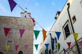 Edificio in pietra con campanile a vela e campana, sormontato dalla stella di David, accanto a un palazzo bianco, con festoni di bandierine colorate tesi sulla strada