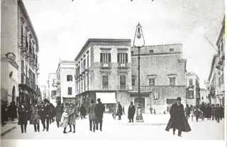 Fotografia d'epoca di un incrocio cittadino con palazzi, passanti e un alto lampione al centro
