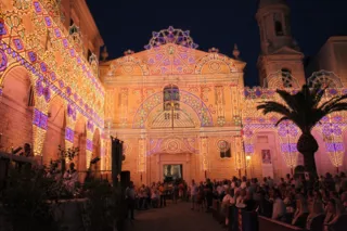 Grande facciata di luminarie colorate allestita davanti alla chiesa per la festa della Madonna del Carmine, con folla nella piazza