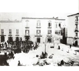 Fotografia d'epoca di una piazza con venditori e banchi del mercato davanti a palazzi ottocenteschi, con un lampione al centro