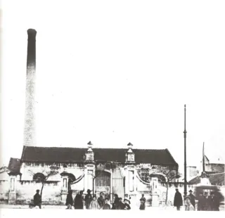 Fotografia d'epoca di un edificio industriale con alta ciminiera in mattoni e cancello ornato, con alcune persone davanti