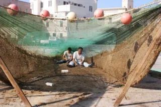 Due uomini seduti a terra riparano una grande rete da pesca stesa con galleggianti, con edifici sullo sfondo