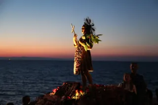 Statua lignea di San Nicola Pellegrino con croce e fronde, adorna di fiori, ripresa al tramonto con il mare e la linea di costa sullo sfondo