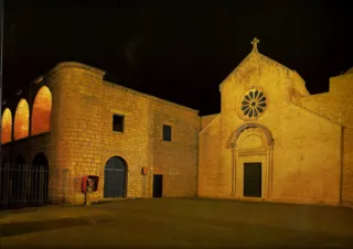 Facciata in pietra della chiesa con rosone e portale, accanto all'edificio del monastero con loggia ad archi, in illuminazione notturna