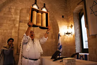 Un uomo con il tallit solleva il rotolo della Torah in un ambiente in pietra con bandiere d'Israele e stella di David