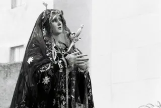 Statua della Madonna Addolorata con manto scuro ricamato, aureola e spada nel petto, portata in processione davanti a una facciata chiara