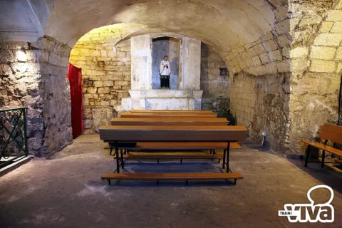 Ambiente sotterraneo con volta in pietra, panche di legno e un altare con una piccola statua