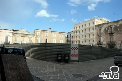 Piazza Campo dei Longobardi chiusa da una recinzione di cantiere in lamiera, con i palazzi della piazza sullo sfondo