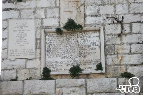 La facciata con la lapide latina, una lapide di marmo a sinistra, uno stemma in alto e un ferro scuro a destra