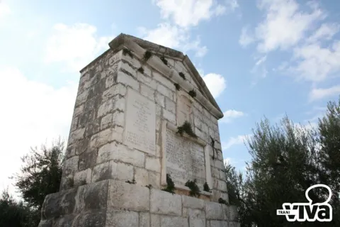 Il monumento di tre quarti visto dal basso: blocchi di pietra, frontone e lapidi con ciuffi d’erba
