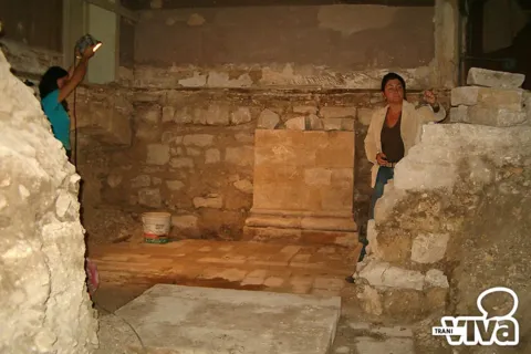 Scavo all'interno dell'edificio: pavimento di piccole pietre e un blocco squadrato, con due donne che fanno luce
