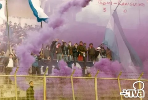 Tifosi sugli spalti fra fumogeni viola e bandiere biancazzurre; sul muro la scritta di una partita finita 3-0