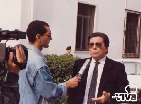 Un giovane cronista con microfono intervista un uomo in giacca e occhiali scuri, ripreso da una telecamera