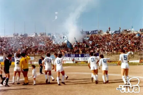 Calciatori in maglia bianca salutano la tribuna gremita, fra fumogeni e bandiere