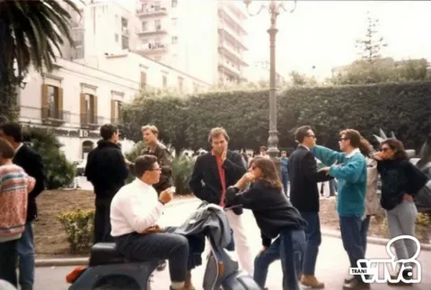 Ragazzi degli anni Ottanta in una piazza di Trani fra palme, siepi e un lampione; uno è seduto su uno scooter