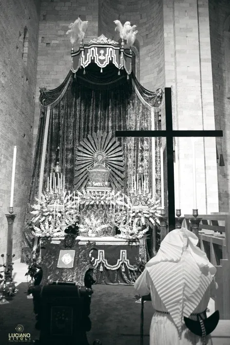 Altare addobbato con baldacchino, fiori e una croce di legno, con un confratello incappucciato in primo piano, in un ambiente in pietra