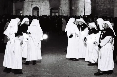 Due file di confratelli con tunica e cappuccio bianchi e mantellina nera, con ceri accesi, in una piazza notturna con la folla sullo sfondo