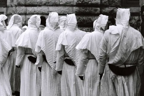 Gruppo di consorelle con veli e cappe bianche e cintura scura, viste di spalle davanti a un muro in conci di pietra
