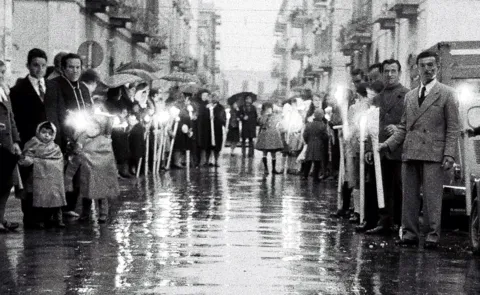 Prospettiva di una via cittadina lucida di pioggia, con due ali di fedeli che reggono ceri accesi in attesa della processione