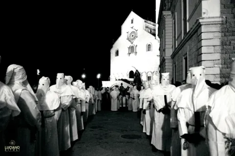 Due file di confratelli incappucciati in bianco e, sullo sfondo, la facciata illuminata della Cattedrale con il rosone, di notte