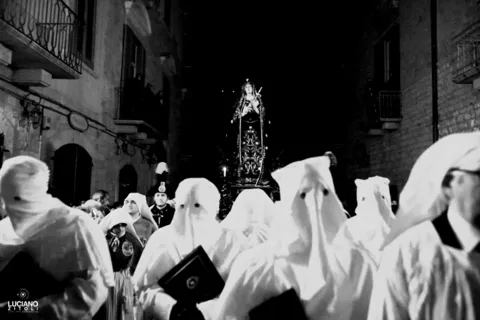 La statua dell'Addolorata avanza in una strada stretta del centro storico, preceduta da confratelli incappucciati