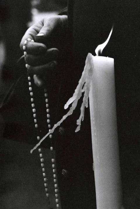Dettaglio in bianco e nero di una mano che tiene un rosario accanto a un cero acceso con la cera che cola