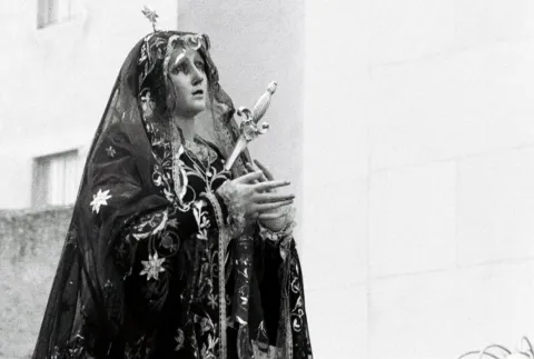 Statua della Madonna Addolorata con manto scuro ricamato, aureola e spada nel petto, portata in processione davanti a una facciata chiara