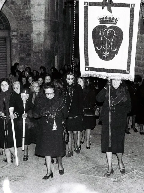 Stendardo con monogramma sormontato da corona portato in processione davanti a un gruppo di donne in abiti neri con ceri