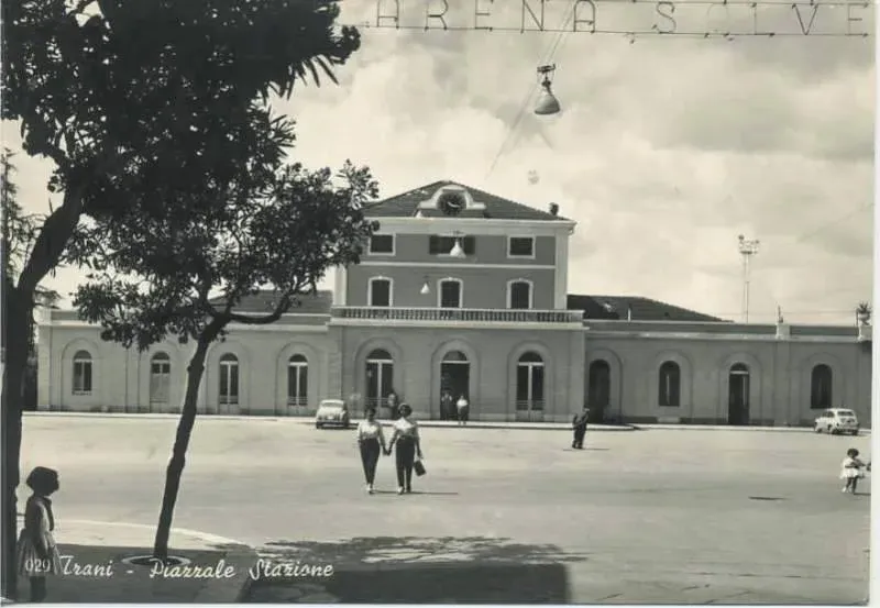 Cartolina in bianco e nero con didascalia 'Trani - Piazzale Stazione': l'edificio della stazione ferroviaria con il piazzale, alcuni passanti e auto d'epoca