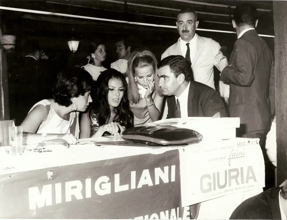 Tavolo della giuria con striscioni 'Mirigliani' e 'Giuria'; due giovani donne e alcuni uomini in giacca conversano chini sul tavolo