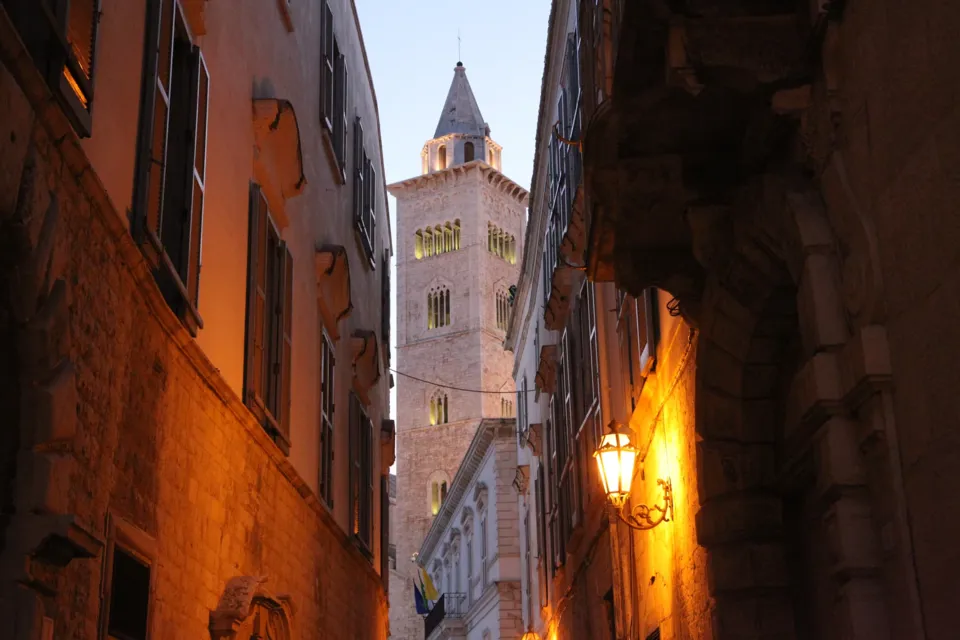 Il campanile della Cattedrale, illuminato all'alba, visto in fondo a una via stretta tra palazzi con lampione acceso