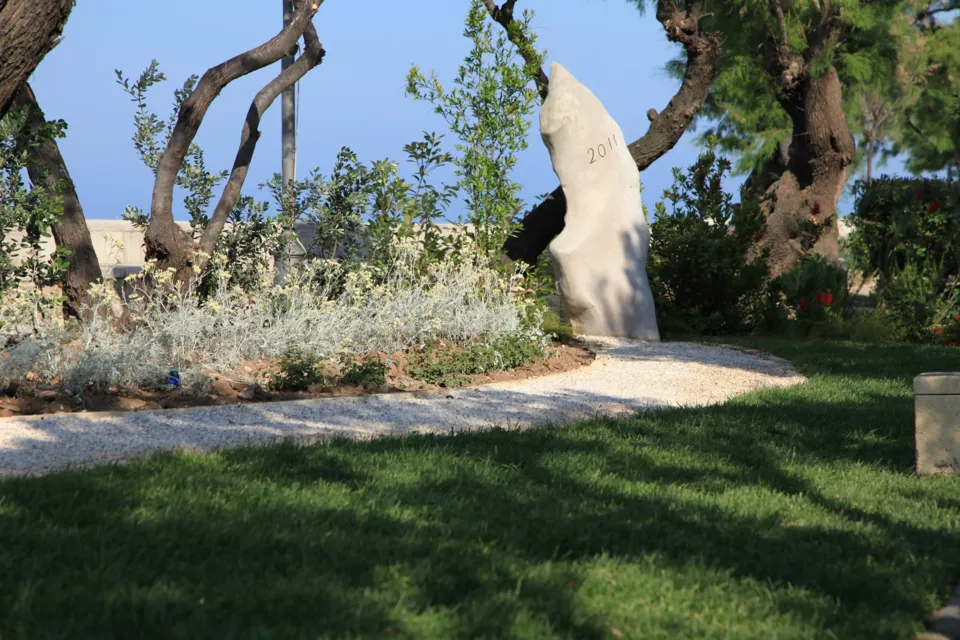 Stele in pietra chiara con l'incisione '2011' in un'aiuola di piante grigie della Villa Comunale, con alberi e il mare sullo sfondo