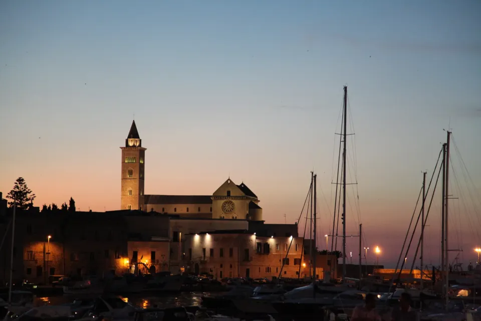 La Cattedrale con il campanile vista dal porto al crepuscolo, con le luci della banchina accese e gli alberi delle barche a vela