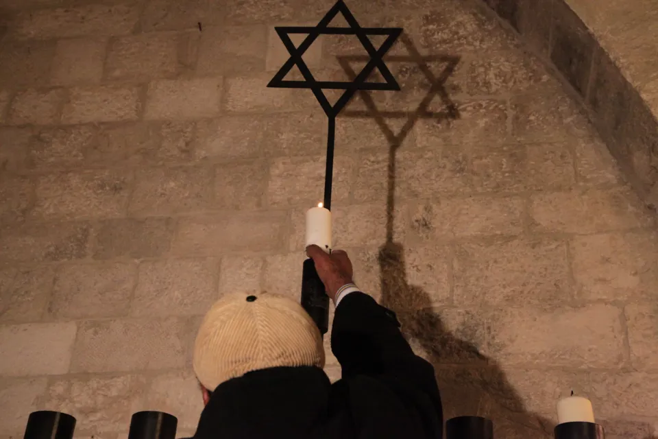 Una mano accende una candela su una grande hanukkiah in ferro sormontata dalla stella di David, contro una parete in pietra