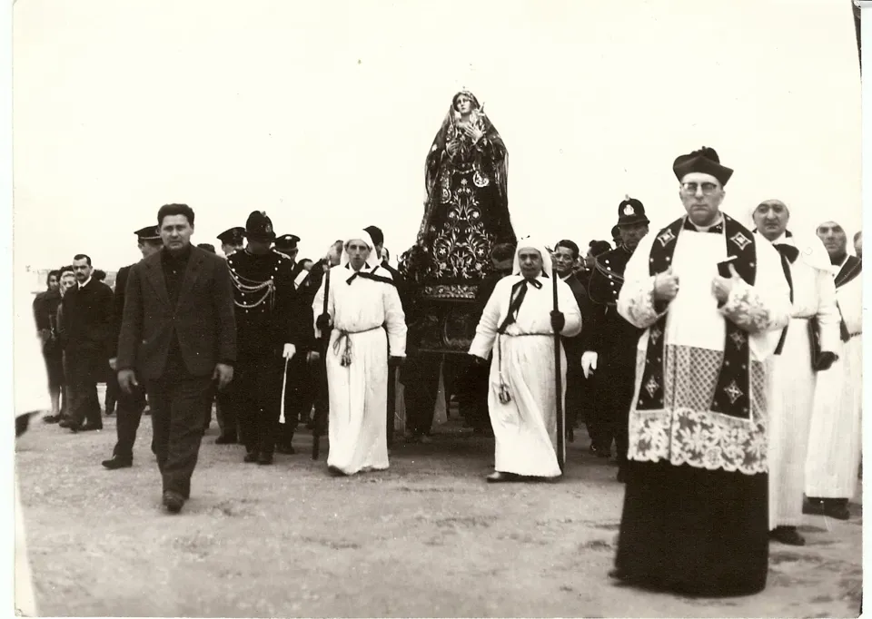 Foto in bianco e nero di una processione con la statua dell'Addolorata, preceduta da un sacerdote in cotta e stola, confratelli in tunica bianca, militari e fedeli