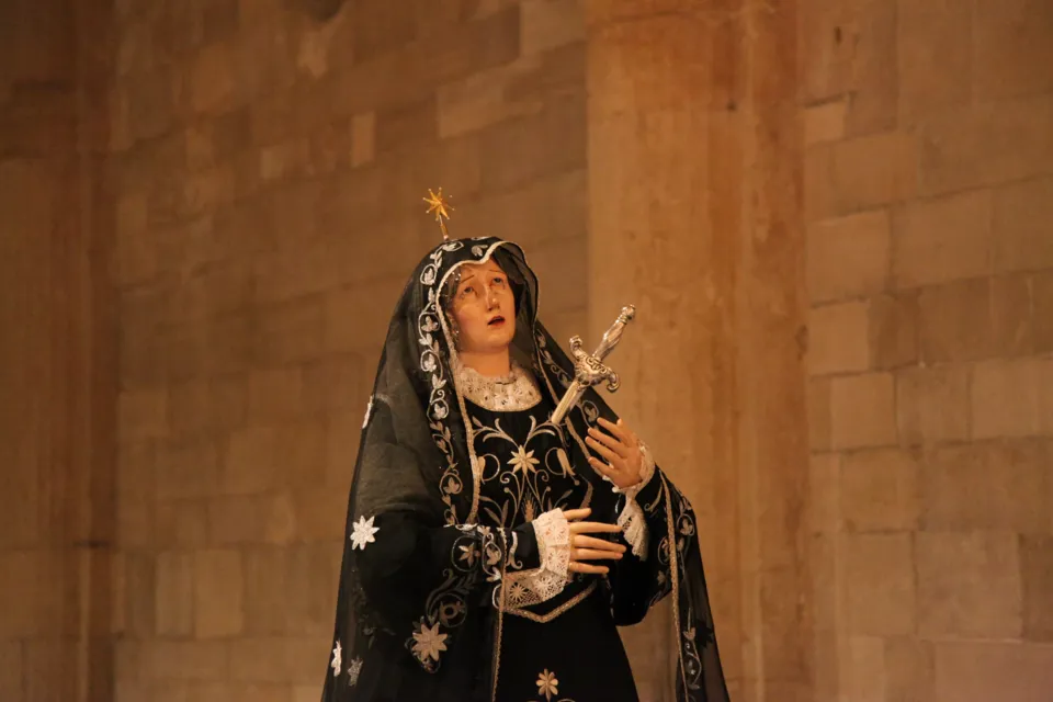 Figura intera della statua dell'Addolorata in manto nero ricamato con stelle, ripresa contro una parete in pietra chiara