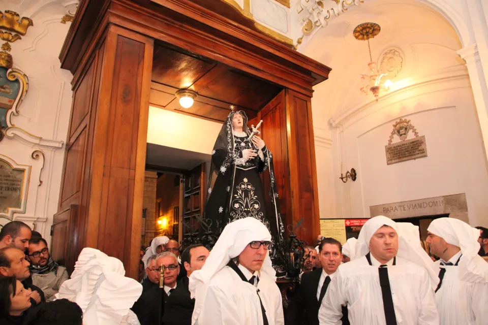 La statua dell'Addolorata in manto nero esce dal portone ligneo di una chiesa con interno chiaro decorato, tra portatori in camice bianco e fedeli