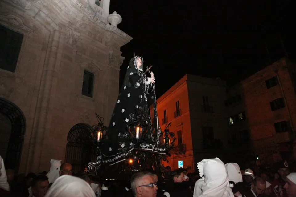 La statua dell'Addolorata, illuminata dai ceri, attraversa una piazza notturna tra fedeli e incappucciati