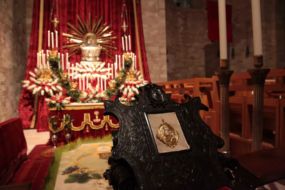 Altare con raggiera e fiori bianchi sotto drappi rossi, ripreso dietro lo schienale di una sedia lignea intagliata con medaglione