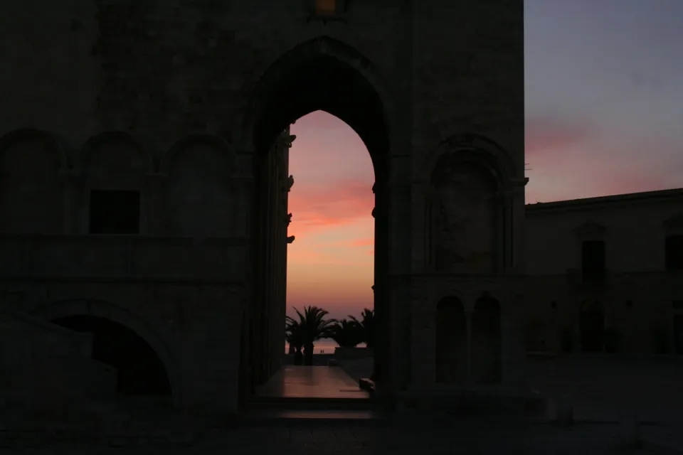 L'arco alla base del campanile della Cattedrale in silhouette, che incornicia il cielo rosa dell'alba e alcune palme