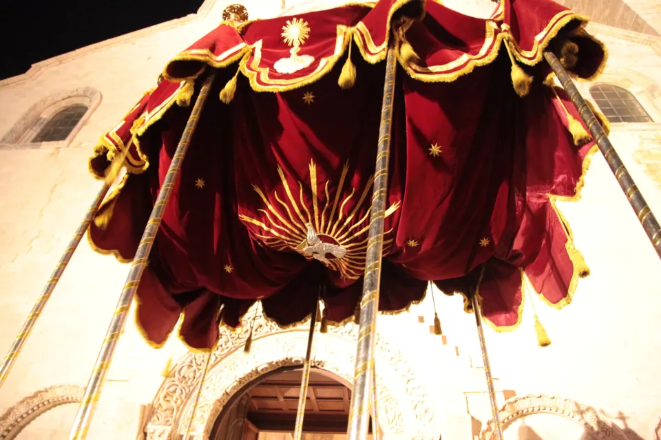 Dal basso, il baldacchino in velluto rosso con frange dorate e raggiera ricamata, sostenuto da aste, davanti alla facciata di una chiesa