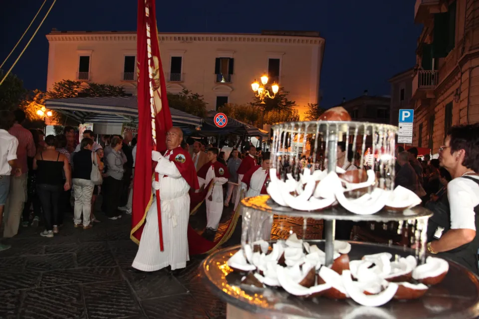 In una piazza di sera, un banco con pezzi di cocco su un'alzata con fontanella in primo piano e un confratello con stendardo rosso tra la folla della festa