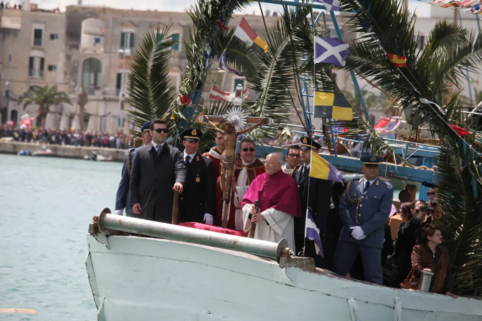 Imbarcazione addobbata con rami di palma e bandiere nel porto, con un prelato, autorità civili e militari e il Crocifisso a bordo