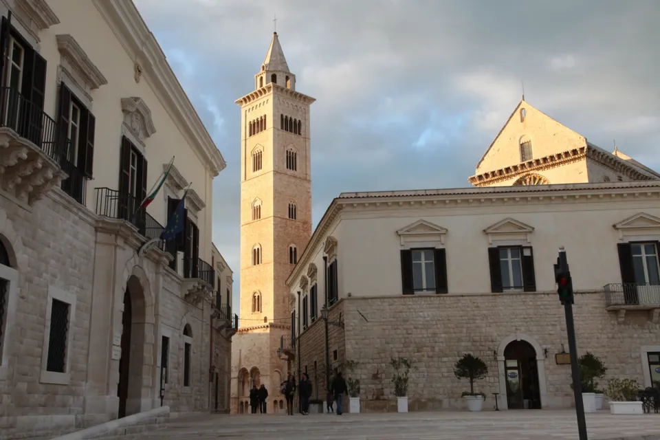Il campanile della Cattedrale visto da una piazza lastricata tra palazzi storici, uno con bandiere, nella luce del pomeriggio invernale