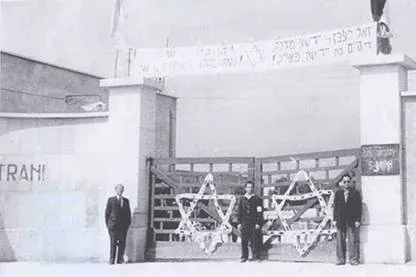 Foto in bianco e nero di tre uomini davanti a un cancello decorato con due grandi stelle di David, sotto uno striscione con scritte in ebraico; sul muro a sinistra la scritta 'Trani'
