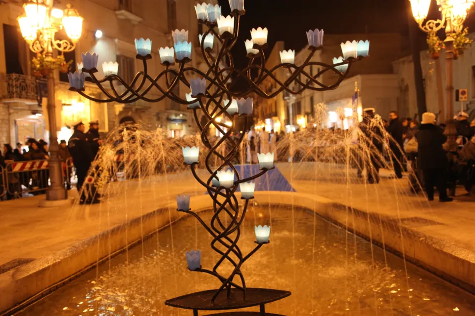 Candelabro in ferro battuto con candele in bicchieri blu al centro della fontana, con la piazza illuminata alle spalle