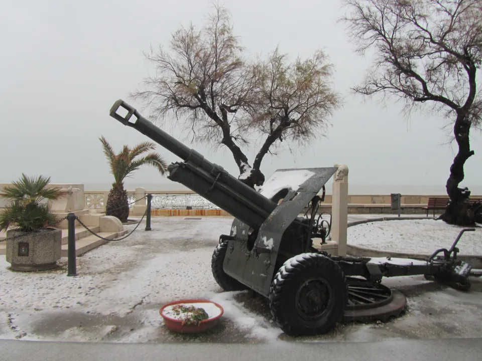 Un obice militare esposto come monumento, coperto di neve, tra alberi spogli e il mare sullo sfondo