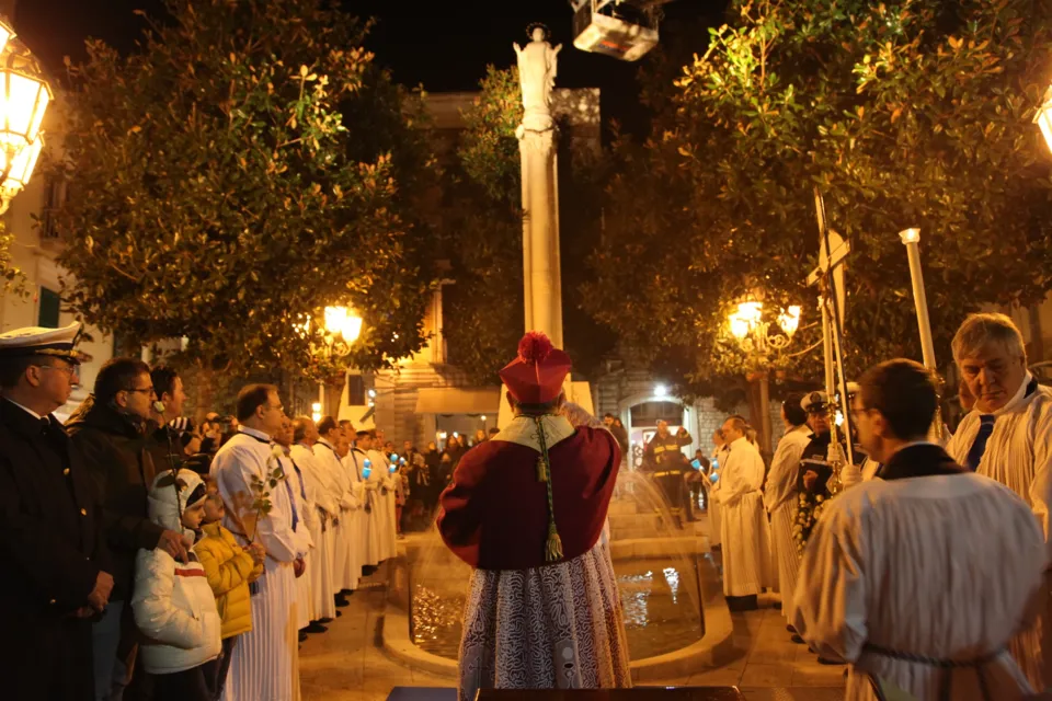Un prelato in mantellina rossa, chierici in cotta bianca e fedeli raccolti ai piedi del monumento all'Immacolata